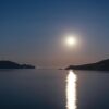 A tranquil night scene with a full moon reflecting on the sea by an island coastline.
