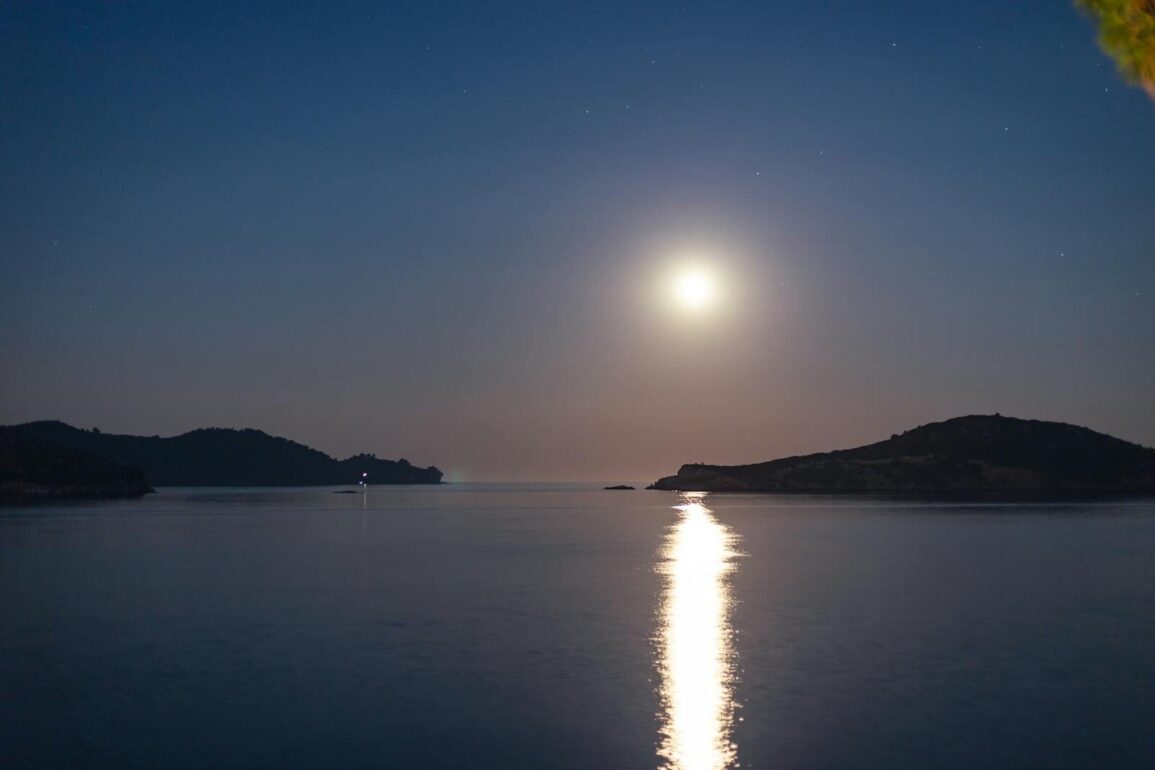 A tranquil night scene with a full moon reflecting on the sea by an island coastline.