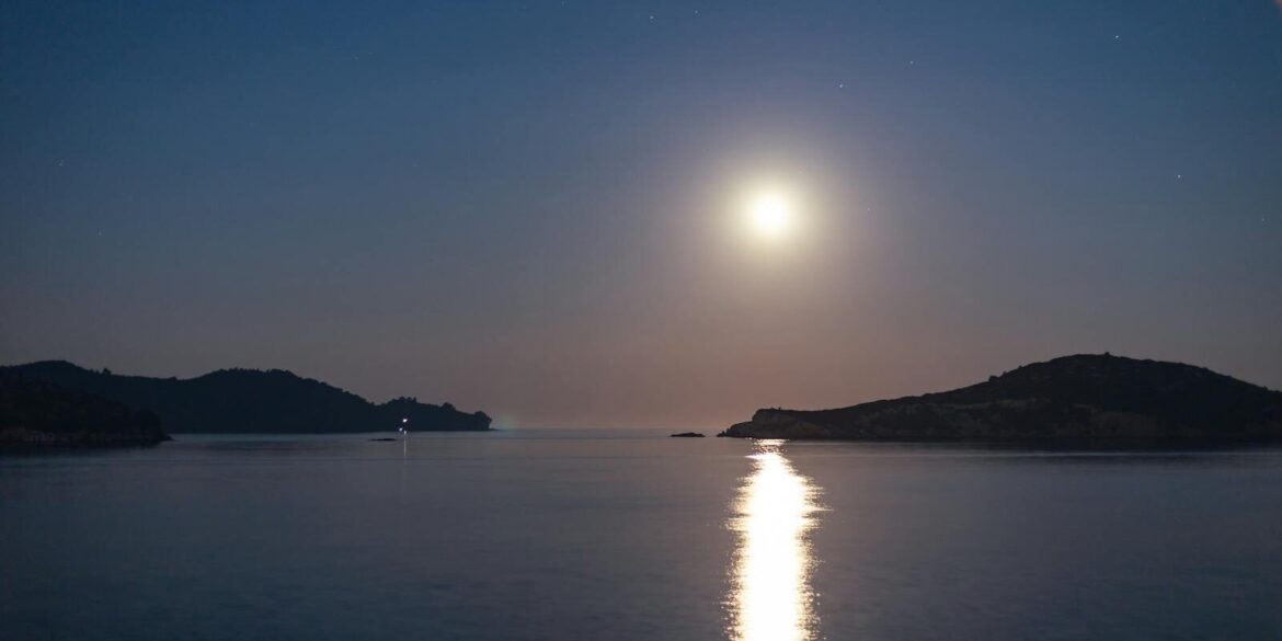 A tranquil night scene with a full moon reflecting on the sea by an island coastline.