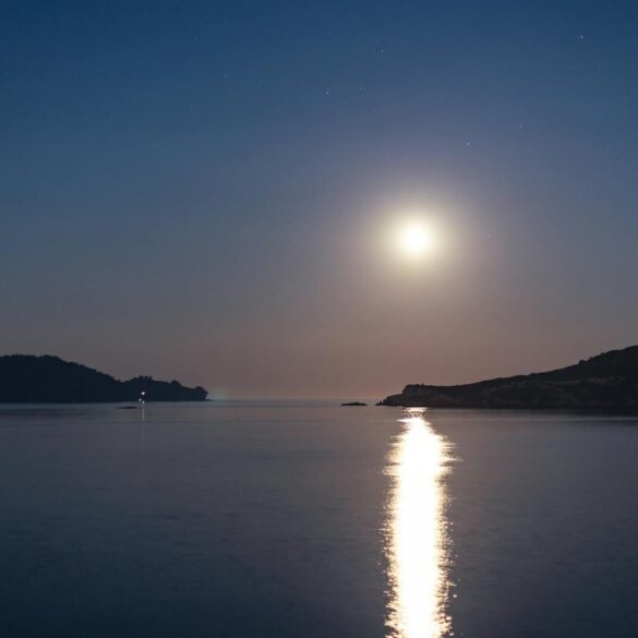 A tranquil night scene with a full moon reflecting on the sea by an island coastline.